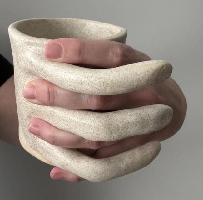 A beige textured mug featuring large ceramic fingers wrapped around the exterior as a handle.