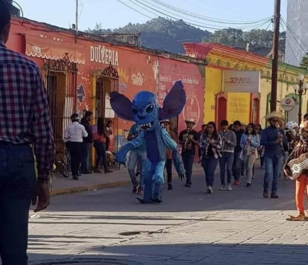 A cursed cosplay of Stitch from Lilo & Stitch walking in a street parade. The costume features a tall, lanky human body and a nightmarish, realistic alien face with sharp teeth and bulging eyes.
