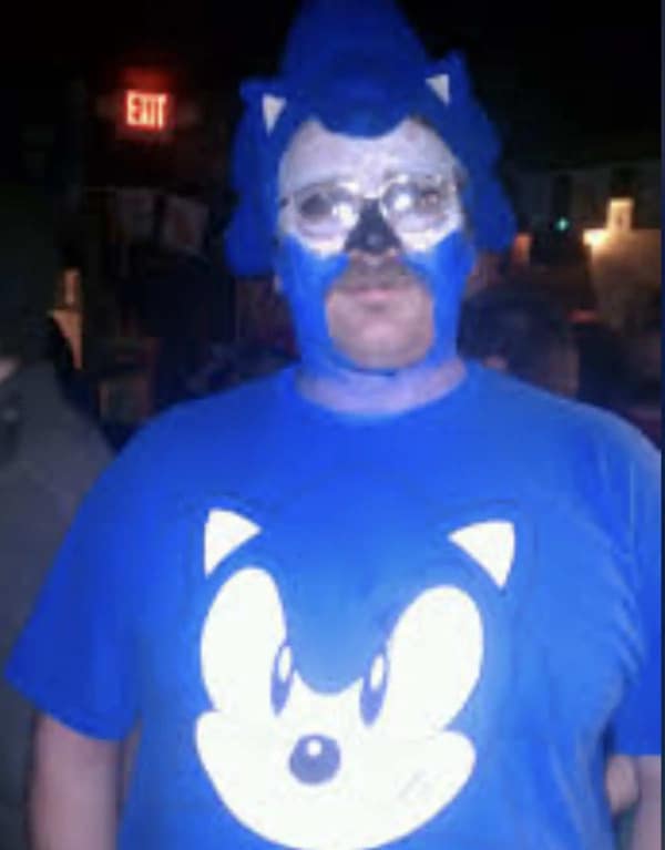 A man with a mustache in a low-effort cursed cosplay of Sonic the Hedgehog, featuring smeared blue face paint and a felt hood that barely covers his head.