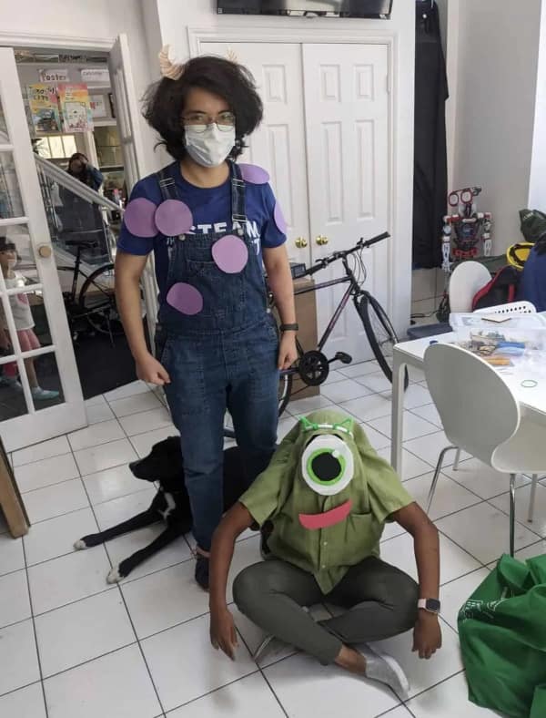 A DIY cursed cosplay duo representing Monsters, Inc., with one person in purple-spotted overalls and another sitting with their head inside a green Mike Wazowski shirt.