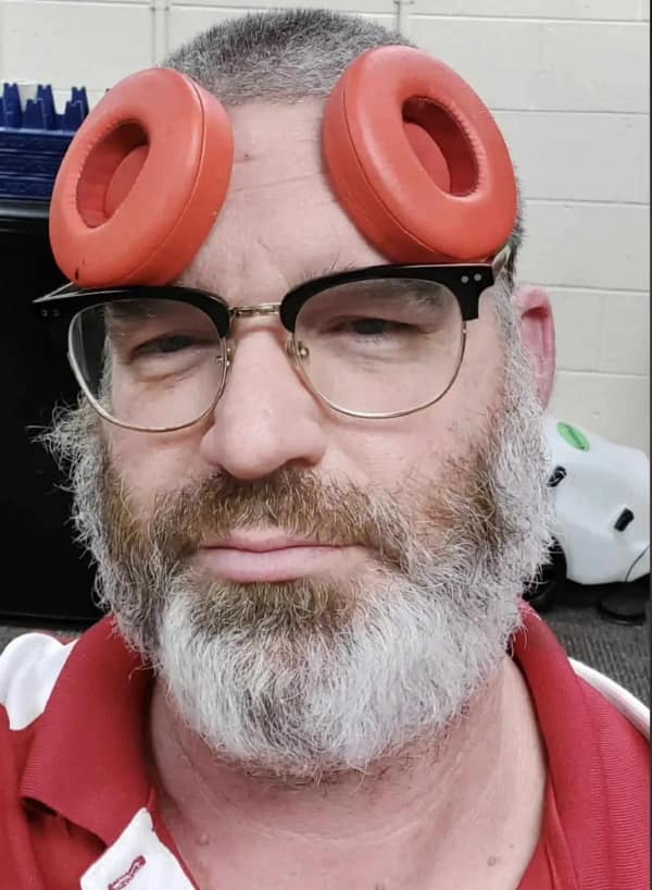 bearded man with glasses performing a low-budget cursed cosplay by sticking two red headphone cushions to his forehead to resemble Mickey Mouse ears or Princess Leia buns.