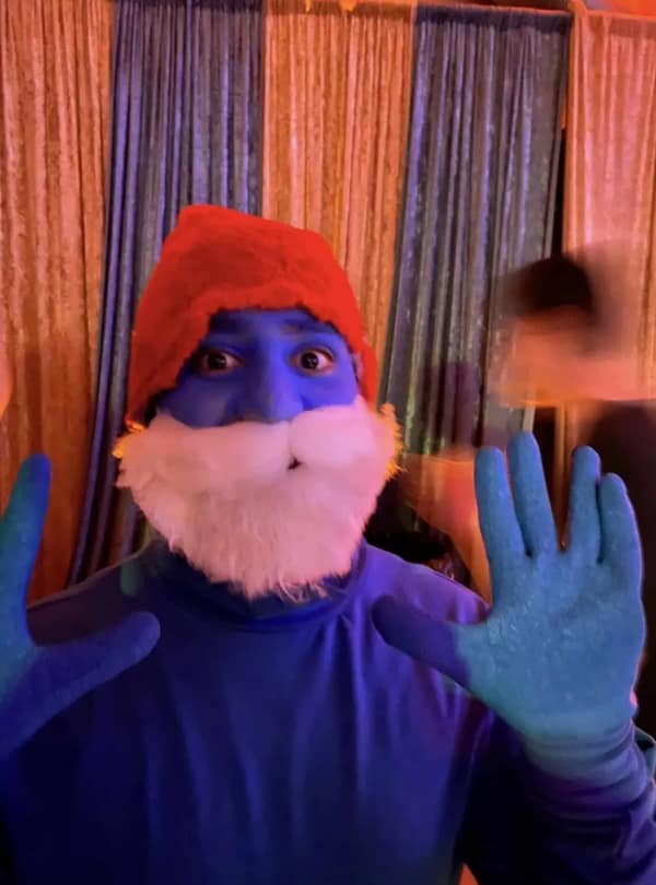 cursed cosplay of Papa Smurf featuring vibrant blue face paint, matching blue gloves, a fluffy white beard, and a red cap, staring directly into the camera with wide eyes.