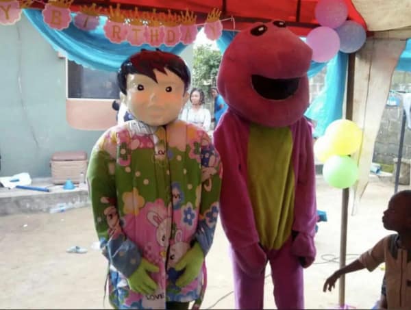 A surreal and cursed cosplay scene at a backyard birthday party featuring a budget, distorted Barney the Dinosaur and a character in a creepy pale mask and floral pajamas.