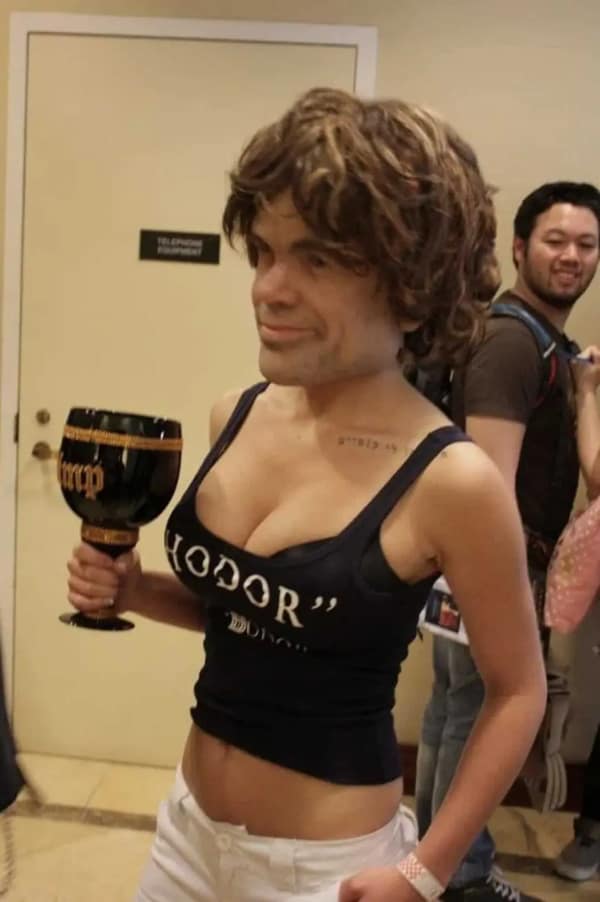 A woman at a convention wearing a realistic, oversized mask of Tyrion Lannister from Game of Thrones while wearing a "Hodor" tank top and holding a golden goblet in a cursed cosplay.