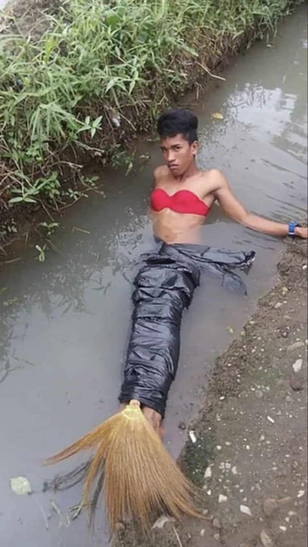 A low-cost cursed cosplay of The Little Mermaid featuring a man sitting in a muddy ditch with a red bra and a tail constructed from black trash bags and a straw broom head.