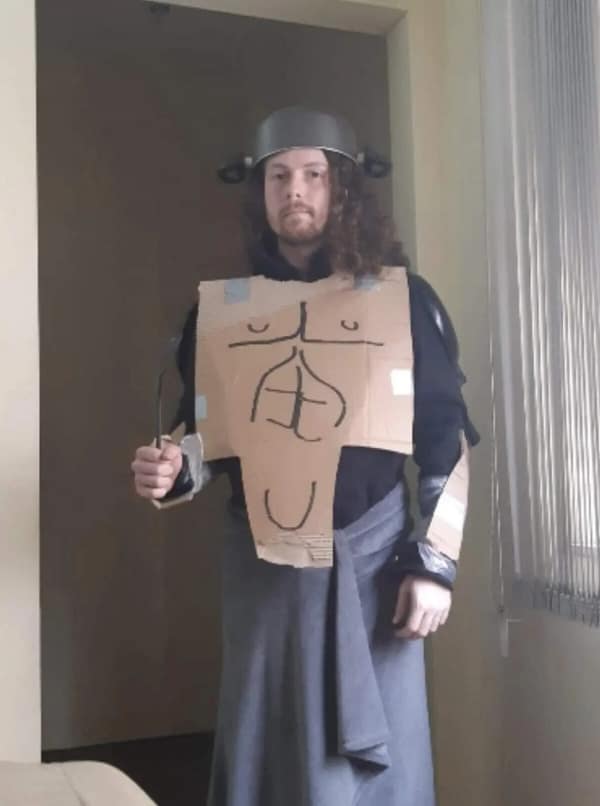 A budget cursed cosplay of a generic fantasy warrior featuring a kitchen pot helmet, a cardboard chest plate with a crudely drawn six-pack, and a grey towel worn as a kilt. Caption: Skyrim belongs to the Nords, but this cardboard belongs to the recycling bin on Tuesday.