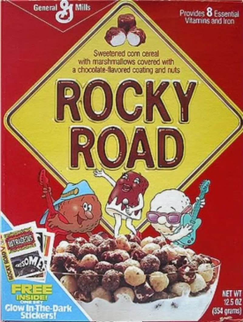 This retro box of General Mills Rocky Road marshmallow-loaded corn cereal brings back memories of discontinued cereals, featuring three cartoon rockstar marshmallow and chocolate characters playing guitars in front of a yellow road sign.