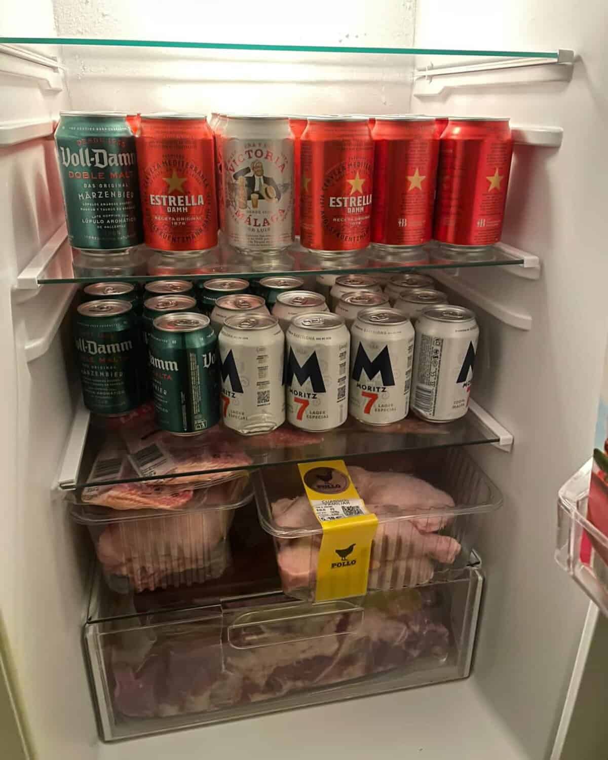 Refrigerator shelves packed tightly with various beer cans and several packs of raw chicken.
