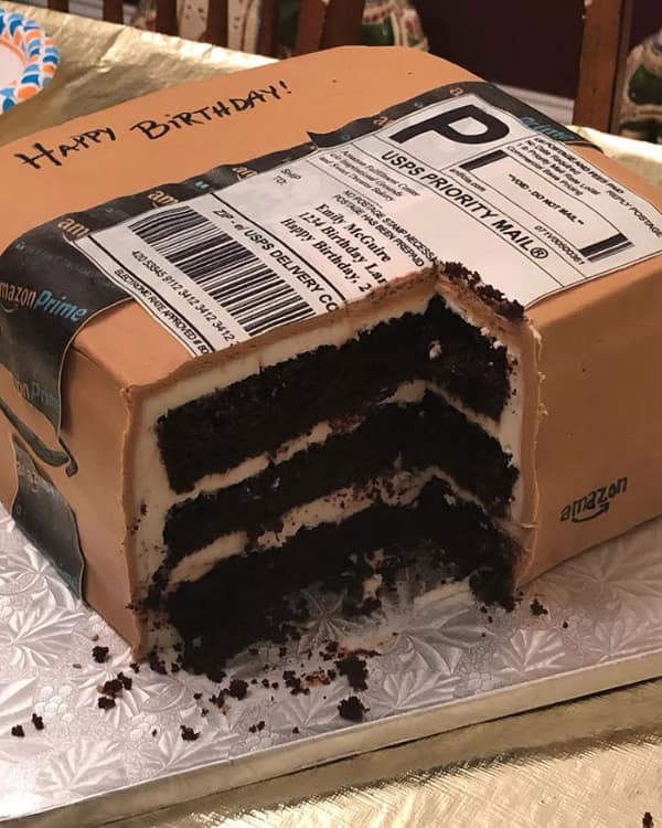 A hyper-realistic custom birthday cake sculpted and frosted to look exactly like a brown cardboard Amazon Prime shipping box with a highly detailed USPS priority mail delivery label.