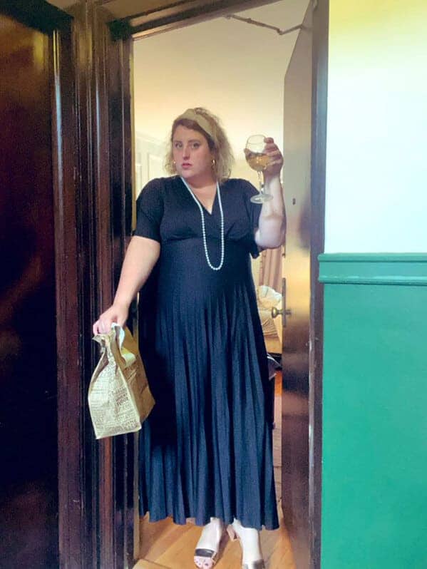 A woman in a fancy black dress and pearls holds a glass of wine and her delivery bag.