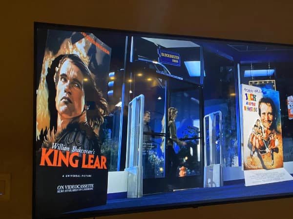 A nostalgic television screenshot captures an alternate-universe parody movie poster inside a retro Blockbuster Video store, featuring Arnold Schwarzenegger starring in "William Shakespeare's KING LEAR" on videocassette and laserdisc.