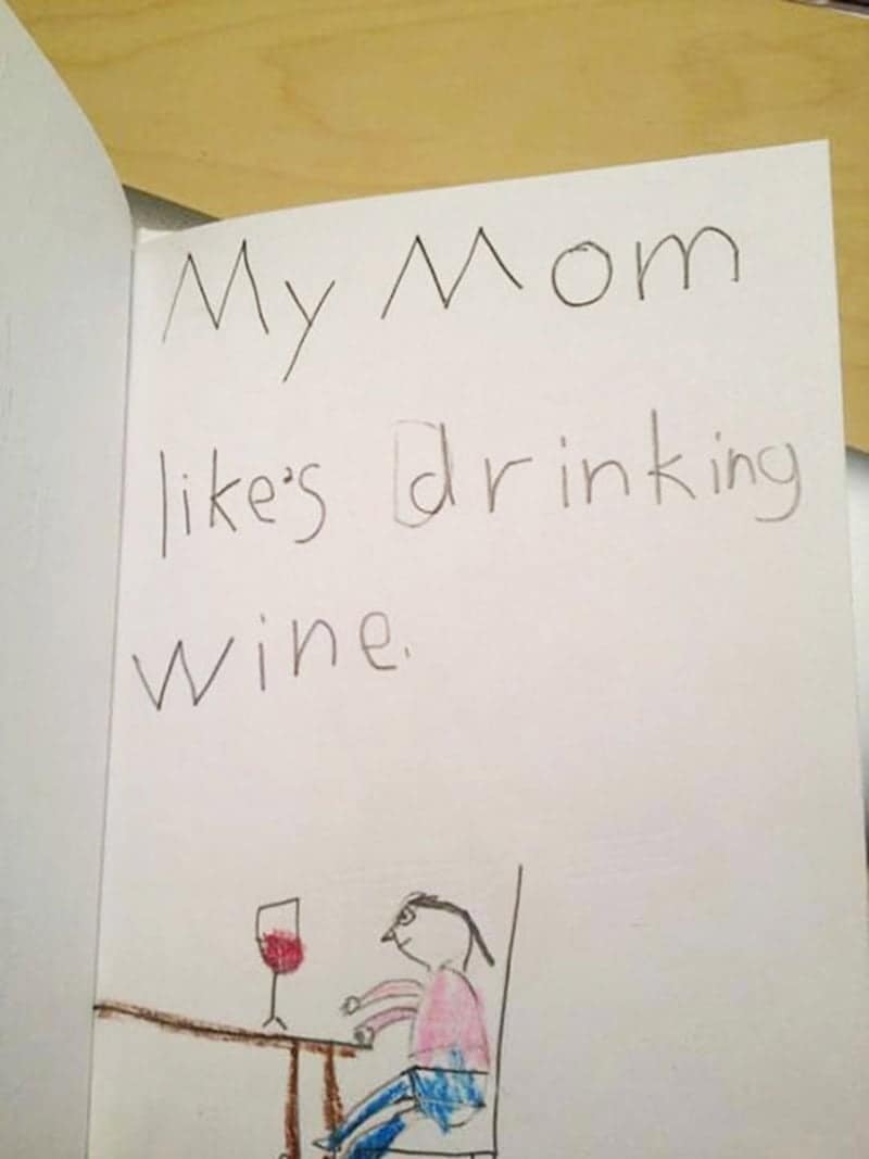 A brutally honest child’s drawing and caption in a school book stating, "My Mom likes drinking wine," featuring a stick-figure mother at a table with a giant red glass.