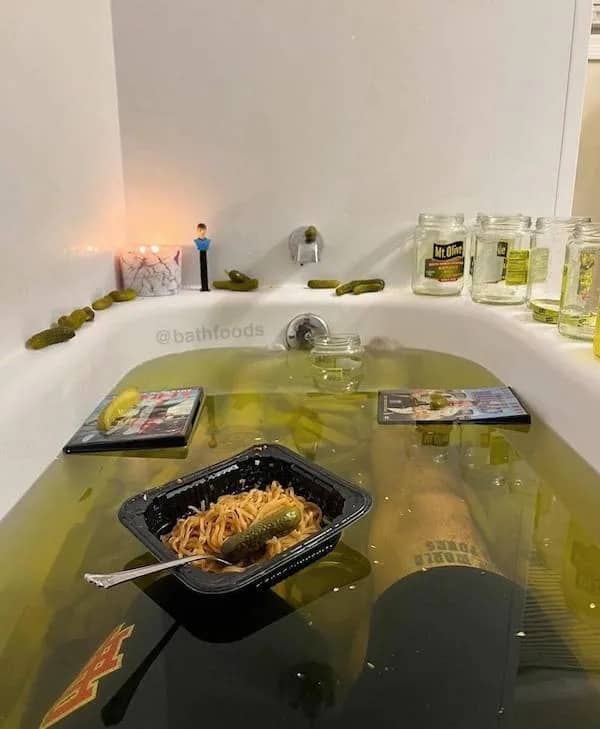 Bizarre bathtub filled with green pickle juice, jars of Mt. Olive pickles, and a bowl of noodles.