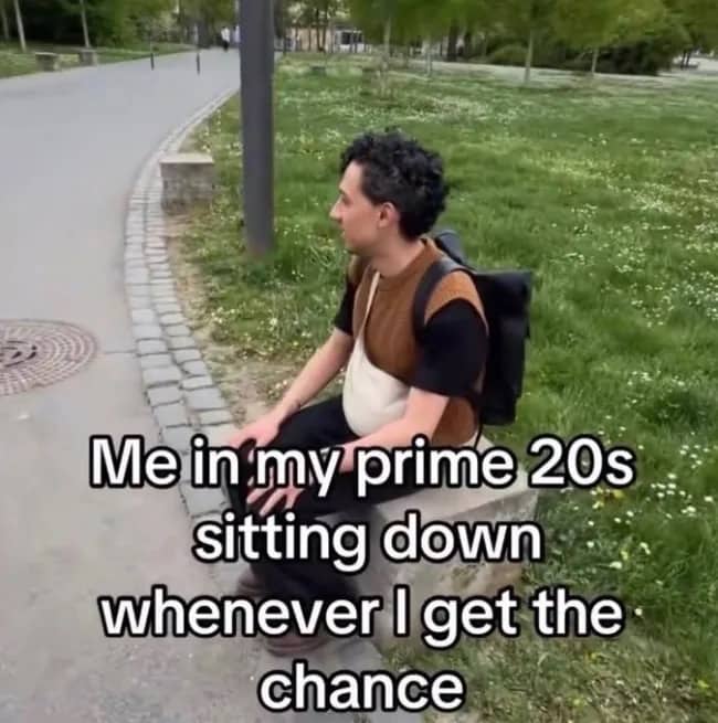 Man with a backpack sitting down on a concrete block in a park looking exhausted.