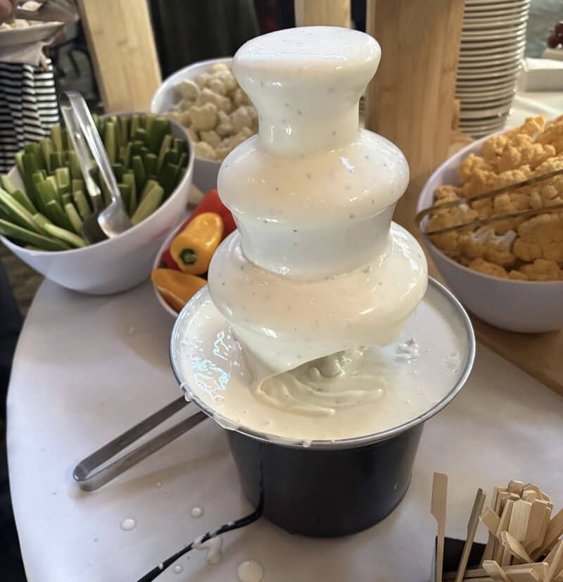 A multi-tiered catering fountain flowing with thick white ranch dressing next to vegetable platters.