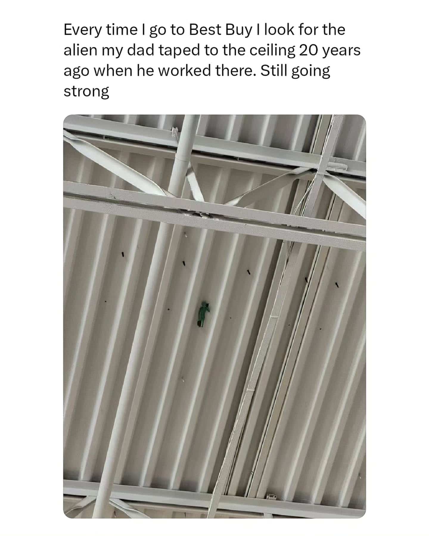 Small green toy alien taped to the corrugated metal ceiling of a large retail store.