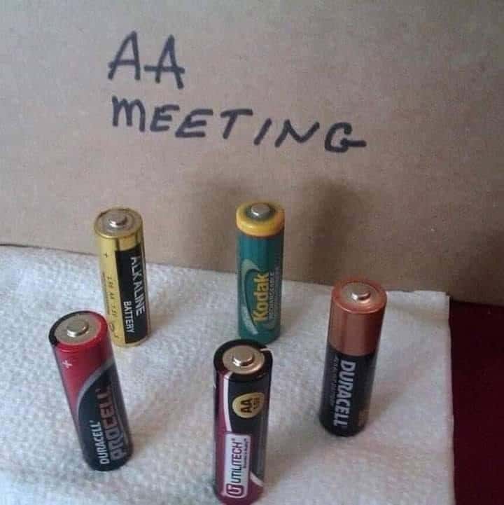 funny pun visual featuring five AA batteries standing in a circle on a paper towel in front of a cardboard sign that reads "AA MEETING."