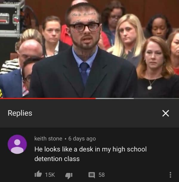 YouTube screenshot of a man with extensive facial and neck tattoos wearing a suit in a courtroom setting. The top reply delivers a sharp funny roast by comparing his ink-covered appearance to a graffiti-strewn desk in a high school detention room.
