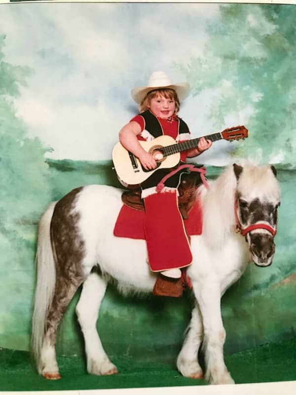 A smiling young girl in cowboy attire holds a guitar while sitting on a pony.