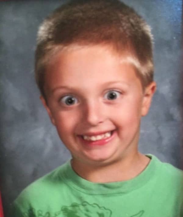 A boy with wide eyes and a manic smile poses against a grey background.
