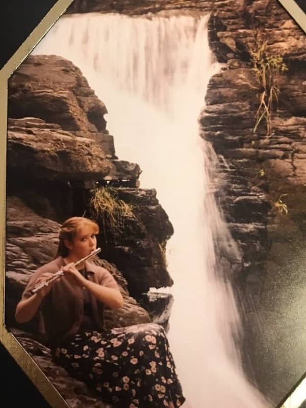 A teenager sits on rocks next to a massive waterfall while playing the flute.