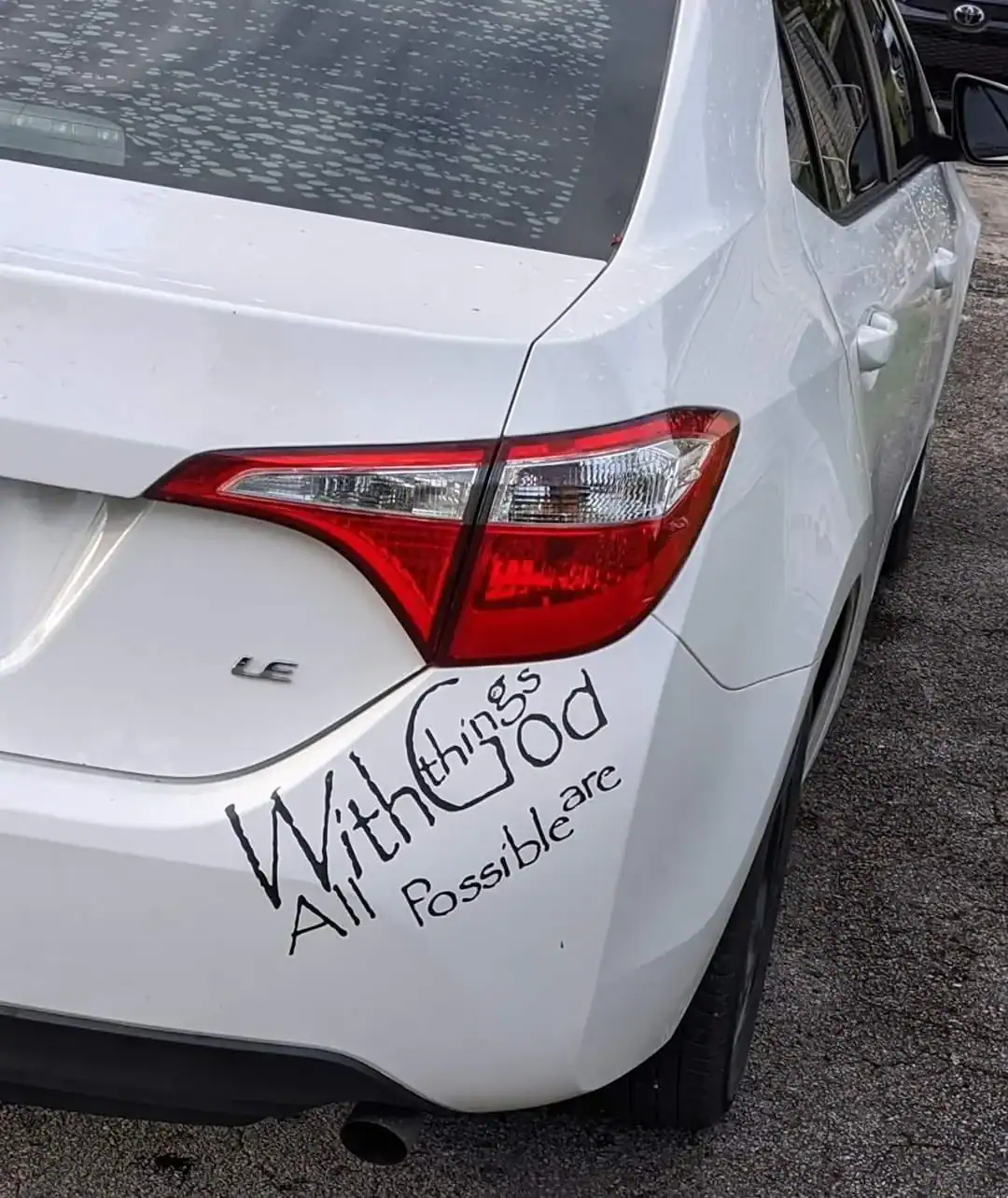A white car with a poorly planned religious quote written in marker on the bumper.