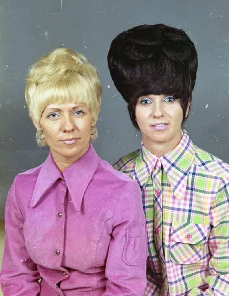Unbelievably high, structural black beehive on a woman with blue eyeshadow, next to a woman with big blonde hair.