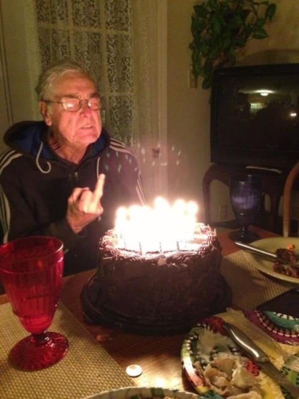 defiant getting old meme showing a grumpy elderly man blowing out birthday candles while holding up a middle finger to the camera.