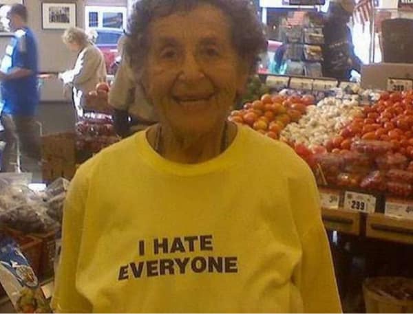 A funny getting old meme showing a smiling elderly woman in a grocery store wearing a bright yellow t-shirt with the bold text "I HATE EVERYONE."
