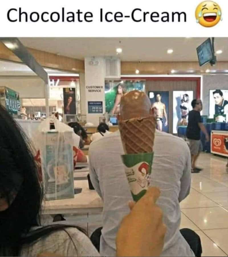 A perspective-bending ice cream meme where a hand holds an empty waffle cone in front of a bald man’s head in a mall, perfectly aligning to make his head look like a scoop of chocolate ice cream.