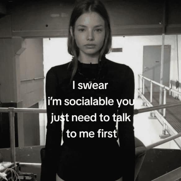 An introvert meme featuring a black-and-white photo of a young woman staring blankly at the camera. White text centered over her reads: "I swear i’m socialable you just need to talk to me first," highlighting the paradox of the passive socializer.