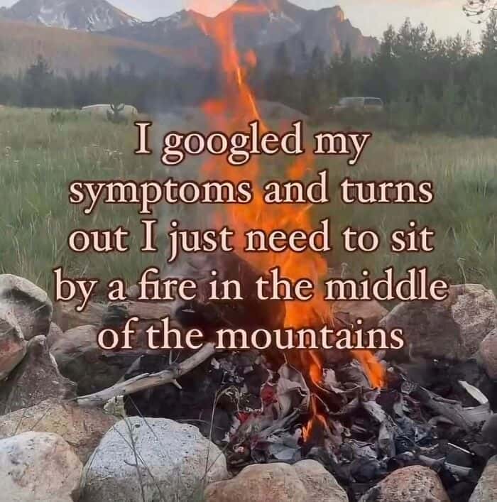 An aesthetic introvert meme showing a cozy campfire crackling in a rocky ring with a vast mountain range in the background at twilight. The text quips: "I googled my symptoms and turns out I just need to sit by a fire in the middle of the mountains."