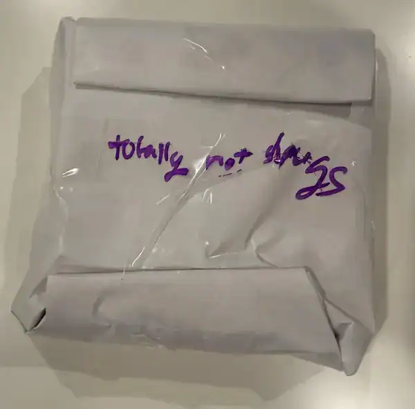 A white rectangular package wrapped in plastic with handwritten text stating totally not drugs.