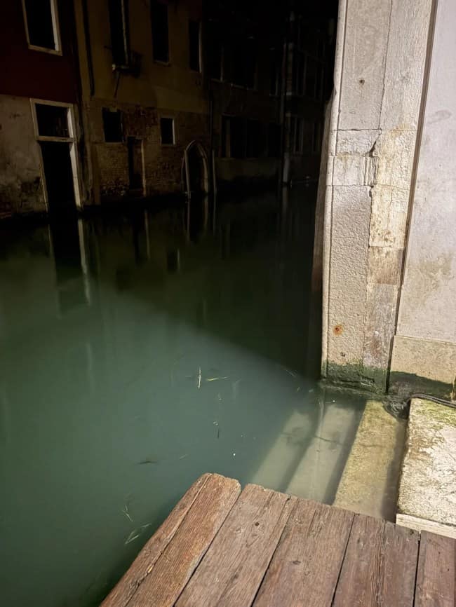 A stone staircase descends straight into the still, murky green water of a dark Venetian canal at night, capturing the submerged secrets of eerie liminal spaces.