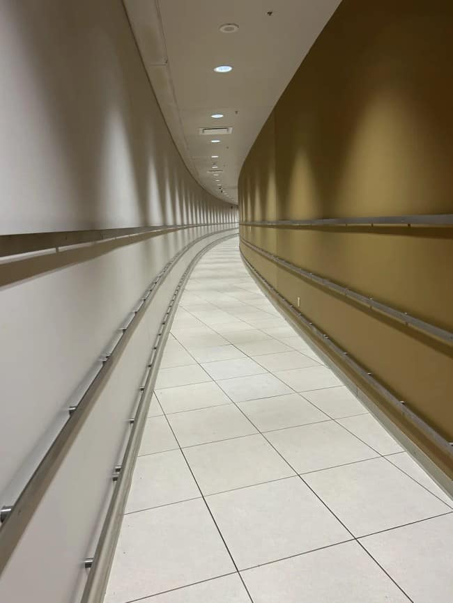 A sterile, brightly lit indoor pedestrian tunnel curves into the distance, featuring crisp white walls on one side and warm gold panels with parallel steel handrails on the other.