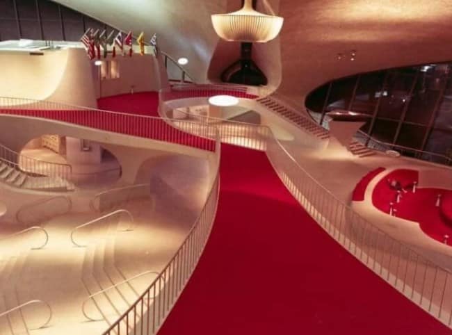 The historic, mid-century modern TWA Flight Center terminal sits completely deserted, emphasizing its organic concrete architecture, multi-level walkways, and sweeping bright red carpet.