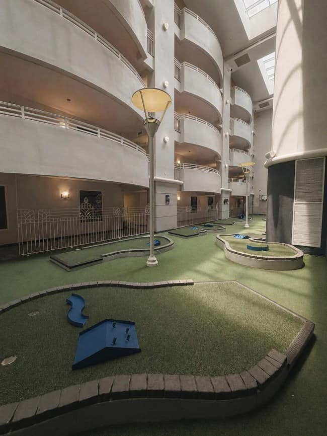 A deserted miniature golf course with green artificial turf sits strangely enclosed inside a multi-story hotel courtyard atrium surrounded by repeating white balconies.