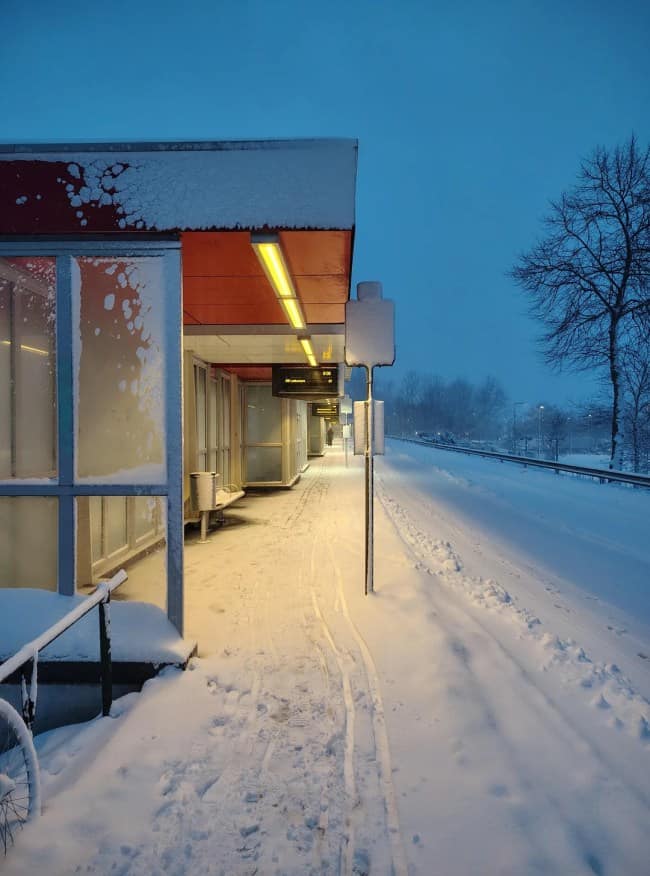 A quiet, snow-covered commuter transit platform at dusk is bathed in the warm, lonely glow of a yellow fluorescent overhang next to empty train tracks.
