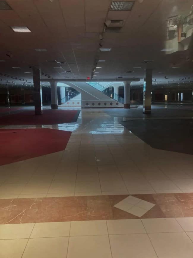 A darkened, abandoned indoor shopping mall corridor features a polished tile floor reflecting a central cross-shaped escalator structure under dim fluorescent utility lighting.