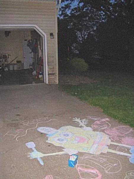 A large, multi-colored sidewalk chalk drawing of SpongeBob SquarePants covers a suburban concrete driveway at night next to an open, dimly lit garage door.
