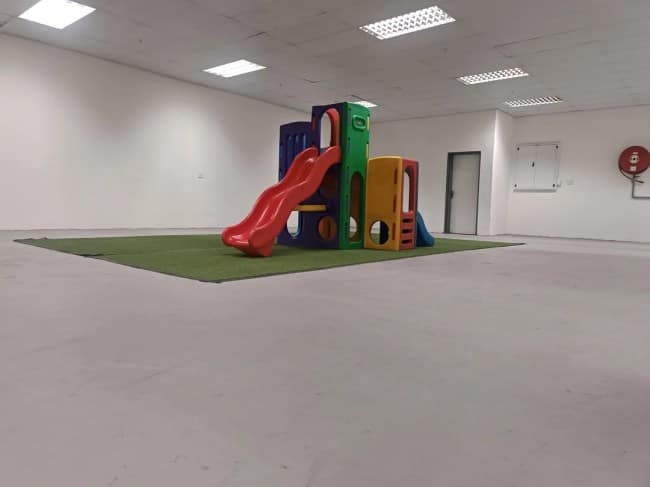 A solitary, multi-colored plastic children's playground slide set sits positioned on a tiny square patch of fake grass inside a massive, empty white room with commercial grid lighting.