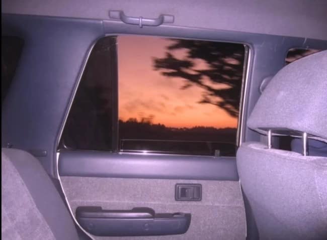 A retro, flash-exposed photo from the backseat of a moving car looks out the passenger window at a blurry, deep orange and purple twilight sunset passing behind silhouetted trees.