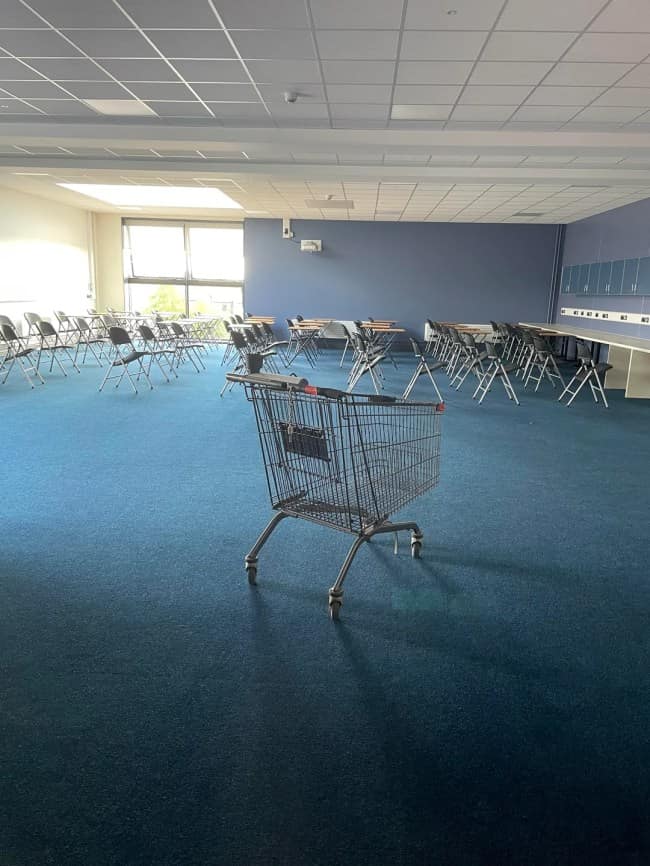 A solitary wire shopping cart sits completely out of place in the center of an empty, blue-carpeted university lecture hall, highlighting the surreal displacements found in eerie liminal spaces.