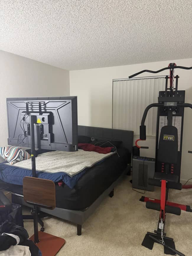 A bedroom layout featuring a television mounted on a stand directly at the foot of the bed beside a home gym machine.
