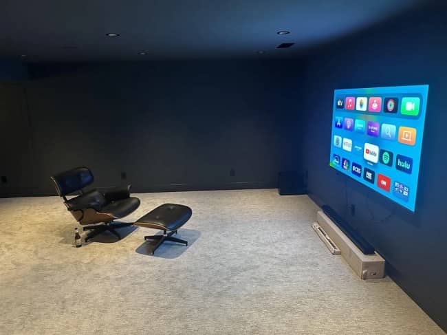 An empty dark blue room containing only a luxury lounge chair, an ottoman, a single bottle, and a television.