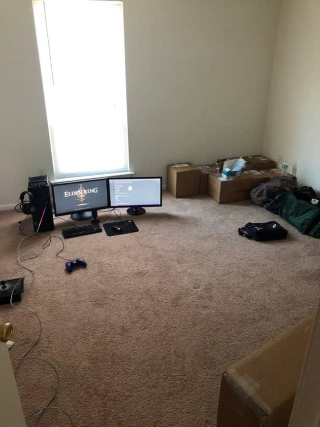 An empty carpeted apartment corner with a dual monitor PC gaming setup resting completely on the floor next to cardboard boxes.