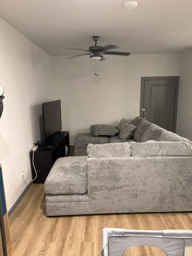 A compact living room completely dominated by a massive gray sectional sofa blocking the main entryway door.