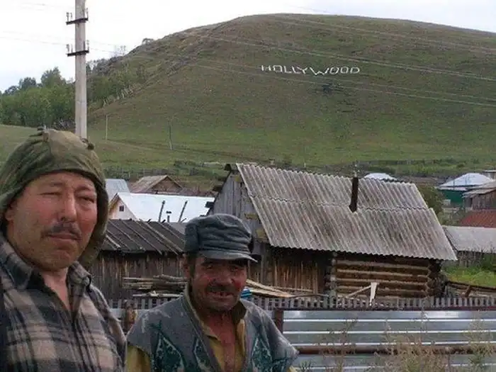 A meanwhile in Russia landscape showing two men in a rural village with a hand-painted, slightly off-model "HOLLYWOOD" sign erected on the barren hillside behind them.