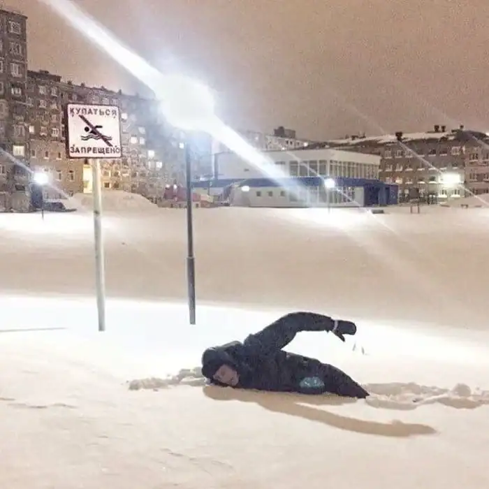 A meanwhile in Russia moment of malicious compliance showing a man "swimming" through a literal snowdrift while lying directly beneath a sign that forbids swimming.