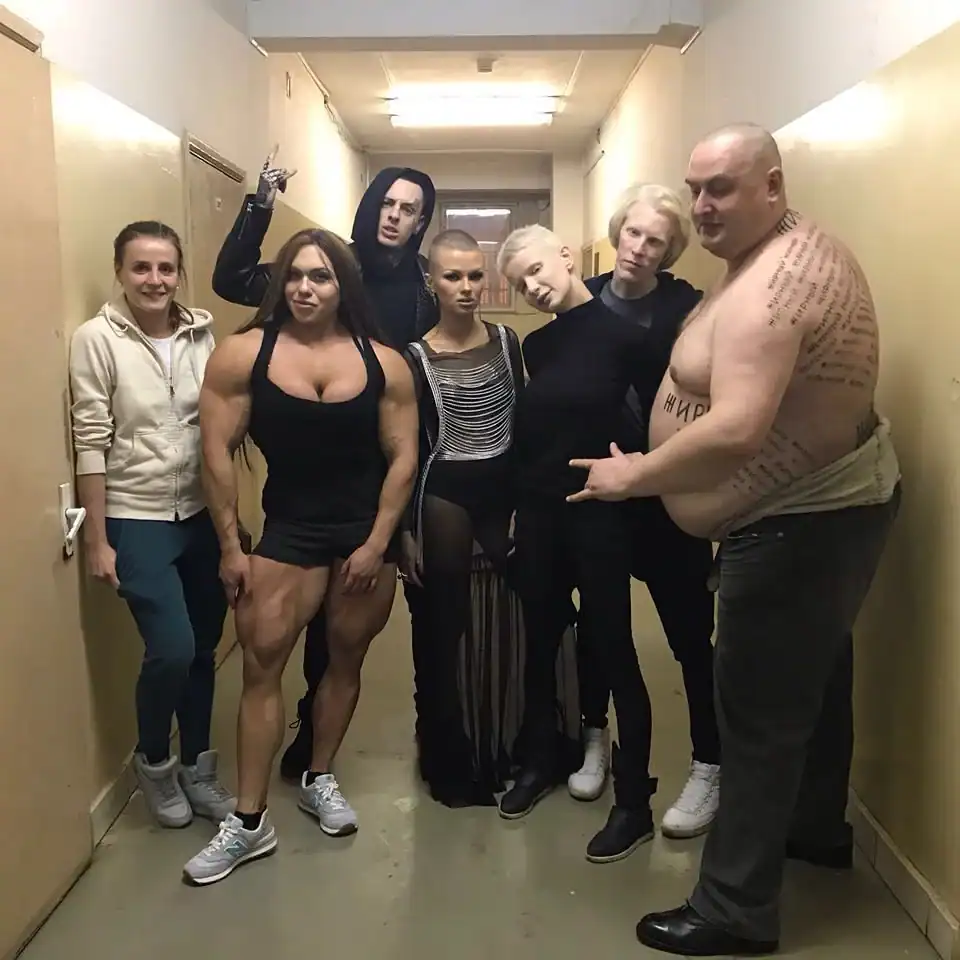 A meanwhile in Russia group photo in a drab hallway featuring a hyper-muscular woman in a tank top, several goth-styled individuals, and a shirtless man with his entire back covered in script tattoos and the word "FAT" in Cyrillic on his stomach.